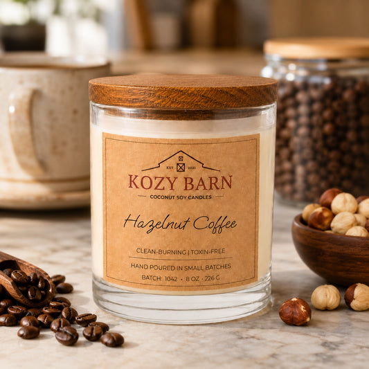 Kozy Barn candle with coconut soy wax and a wooden lid labeled Hazelnut Coffee styled on a marble surface with coffee beans, a bowl of hazelnuts, and a ceramic mug in the background.