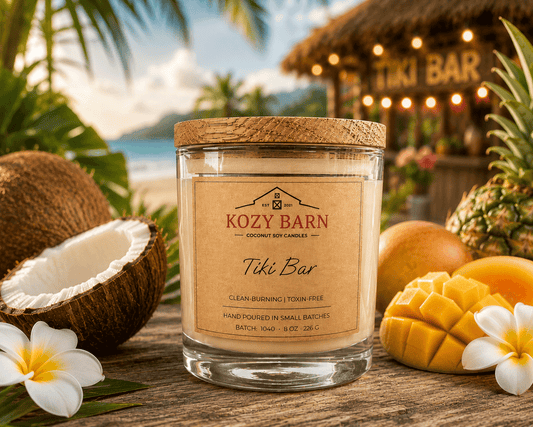 Kozy Barn candle labeled Tiki Bar sitting on wooden table next to pineapple, mango, orange, halved coconut and white flower blossoms, in the background there is a tiki bar and ocean beach.