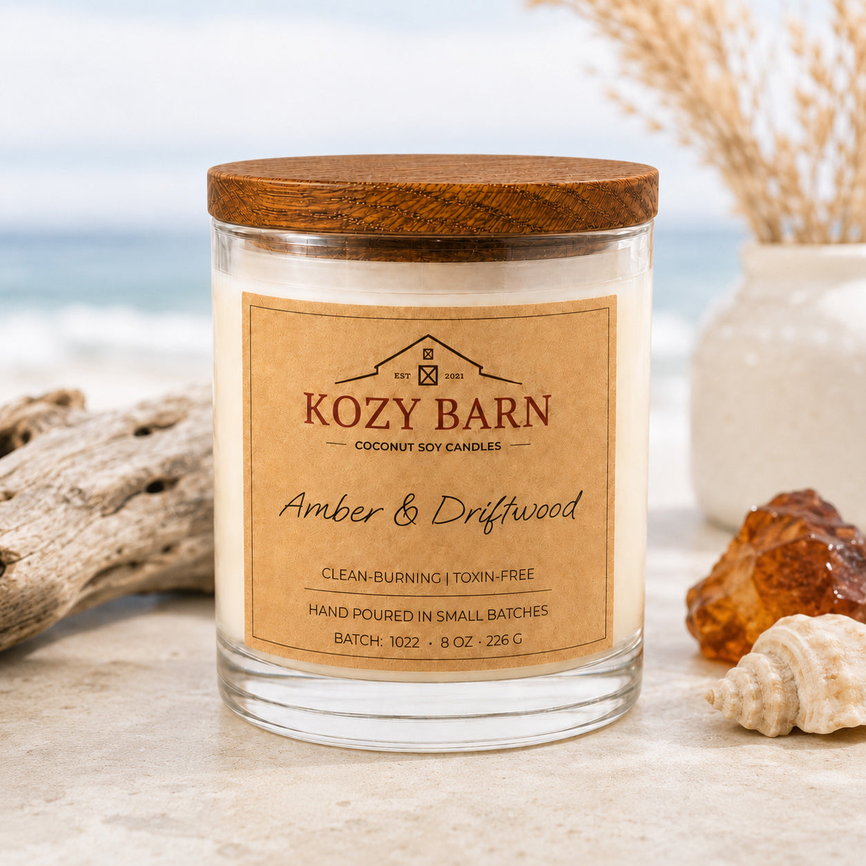 Kozy Barn candle with coconut soy wax and a wood lid labeled Amber & Driftwood set on a stone surface with driftwood, shells, and a blurred beach background.