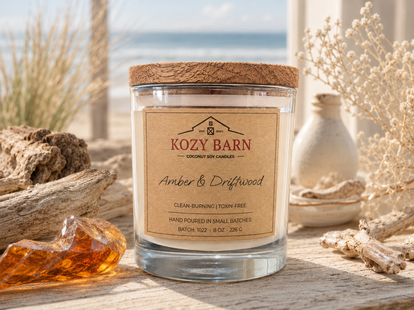 Kozy Barn candle labeled Amber & Driftwood sitting on beach wood next to amber, driftwood and ocean beach in the background.