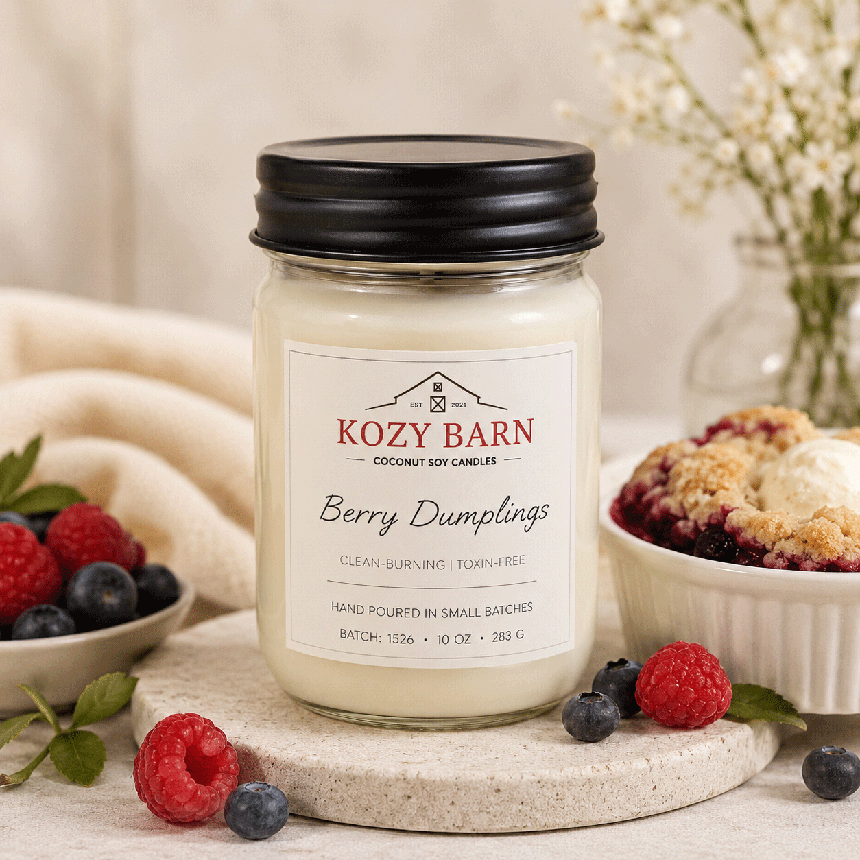 Kozy Barn candle labeled Berry Dumplings sitting next to black raspberry dumplings and a bowl of red raspberries and blueberries.