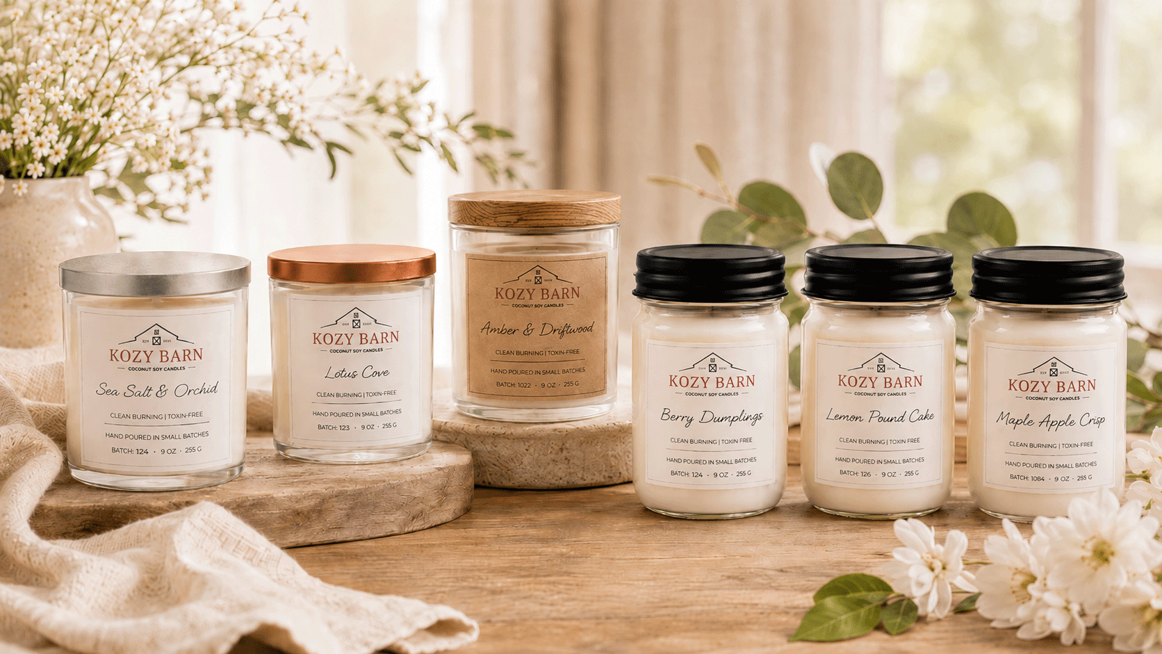 Six jars of Kozy Barn scented candles arranged on a wooden surface with decorative flowers and fabric in a softly lit indoor setting. The candles come in clear glass jars with white wax and lids in silver, copper, wood, and black finishes.