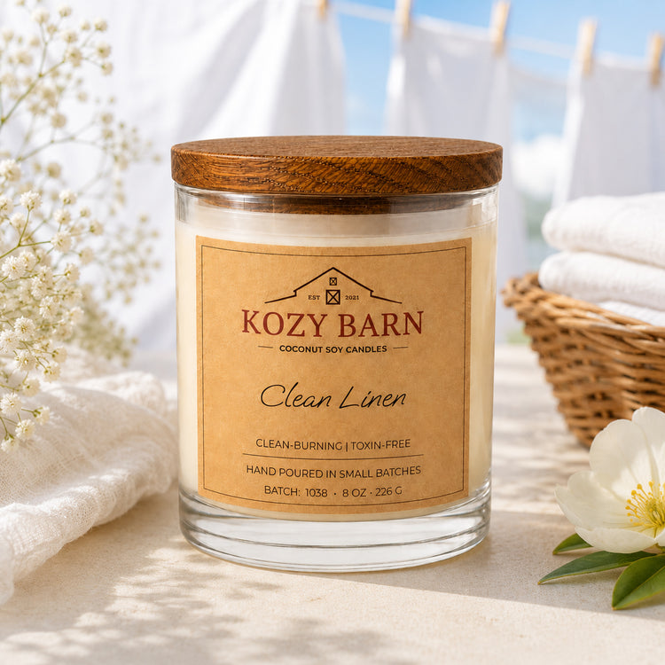 Kozy Barn candle with coconut soy wax and a wooden lid labeled Clean Linen sits on a light surface with soft white towels, a wicker basket, and a white flower in a bright, airy setting.
