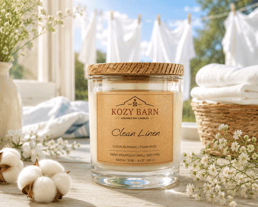 Kozy Barn candle labeled Clean Linen sitting in a window next to cotton balls and basket of clean white towels with hanging white laundry in the background.