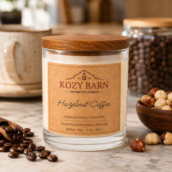 Kozy Barn candle with coconut soy wax and a wooden lid labeled Hazelnut Coffee styled on a marble surface with coffee beans, a bowl of hazelnuts, and a ceramic mug in the background.