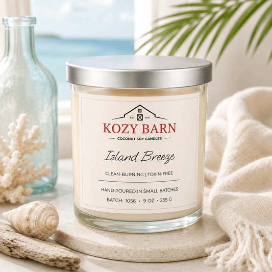 Kozy Barn candle labeled Island Breeze sitting on wooden table next to glass bottle, seashells, and soft cozy blanket background has green fern and window with ocean. 