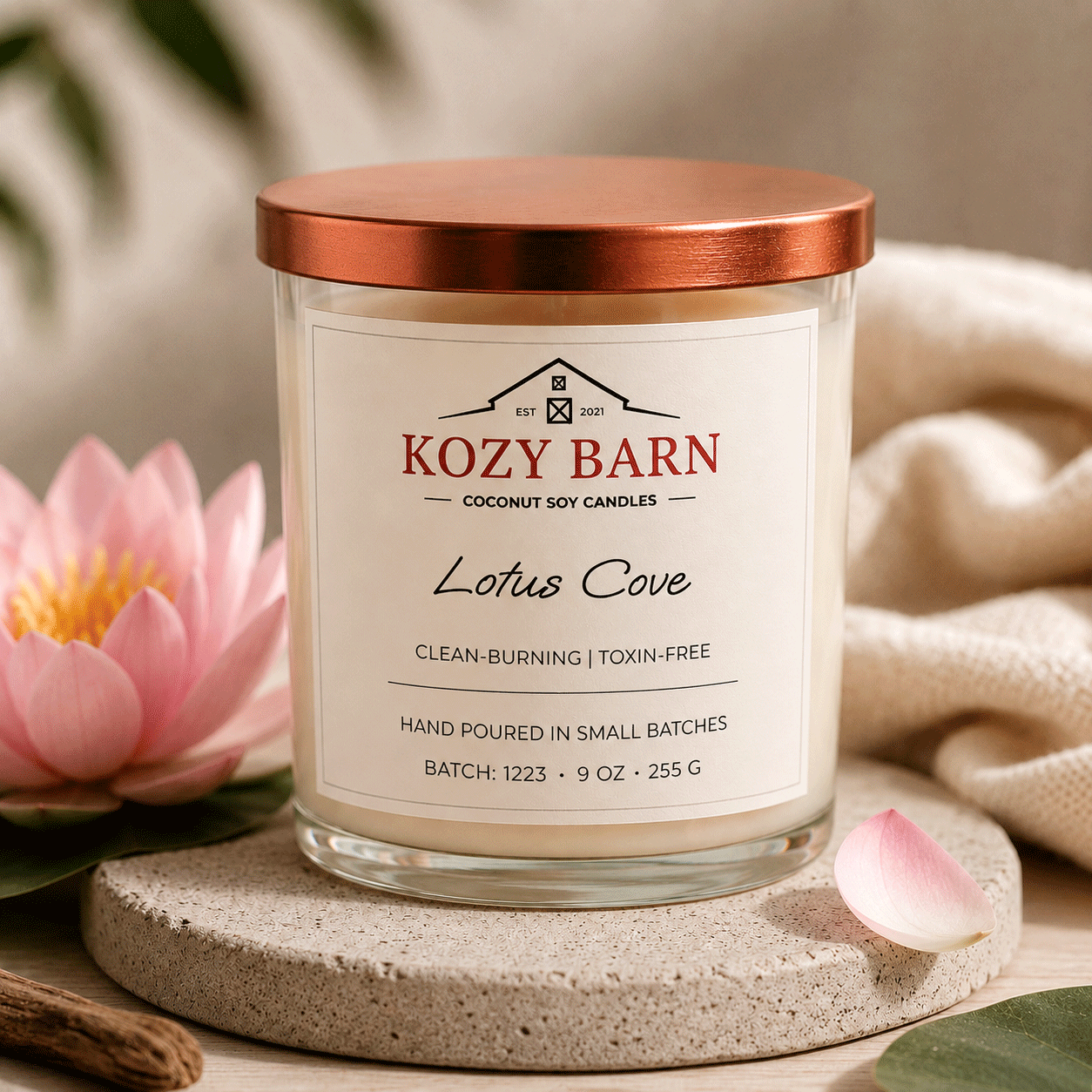 Kozy Barn candle labeled Lotus Cove sitting on table next to pink blossoms and cozy soft blanket. 
