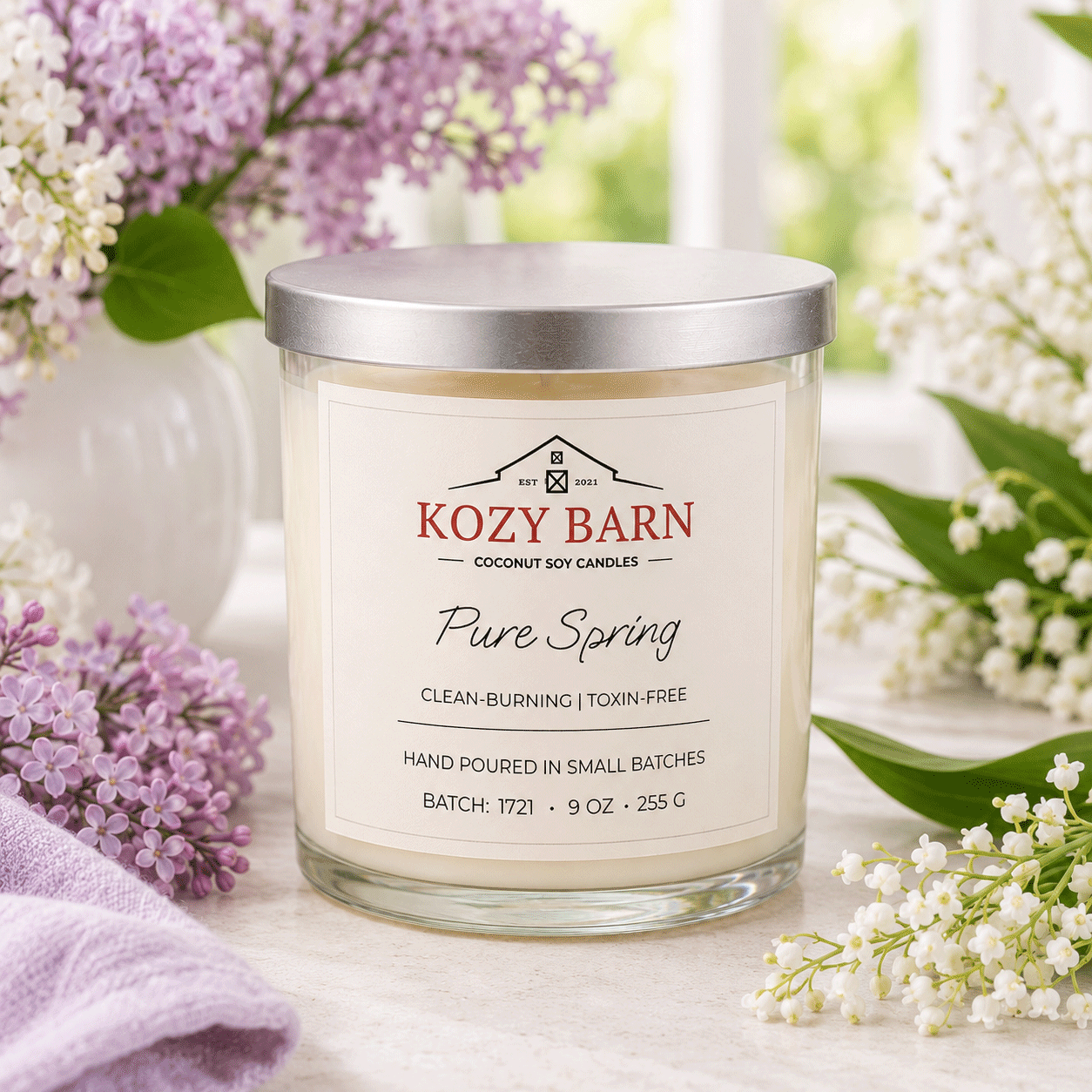 Kozy Barn candle labeled Pure Spring sitting on a table next to baby French Lilac and Lilly of the Valley. 