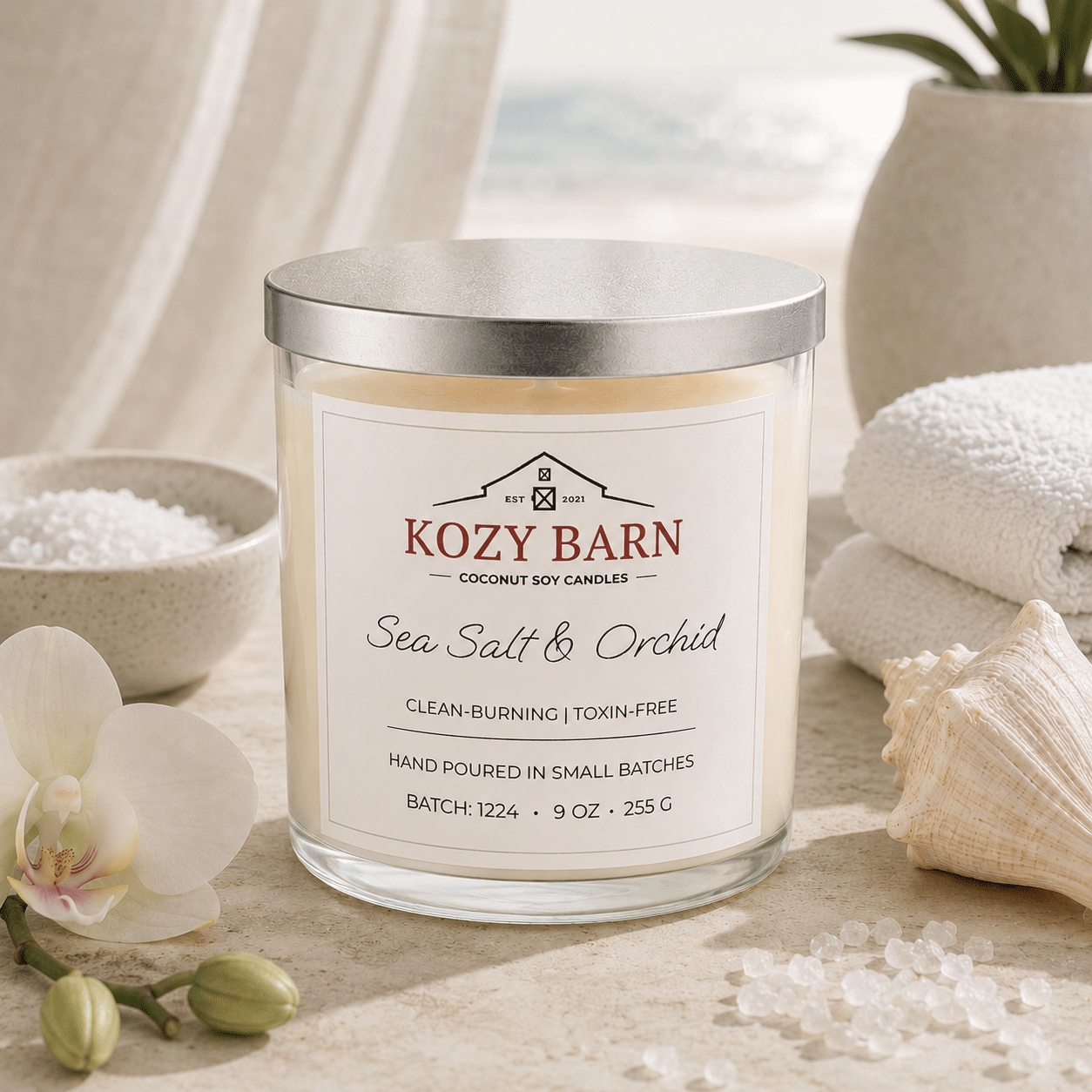 Kozy Barn candle labeled Sea Salt & Orchid sitting a beach table next too bowl of salt, orchid blossoms, sea shell and white spa towels. 
