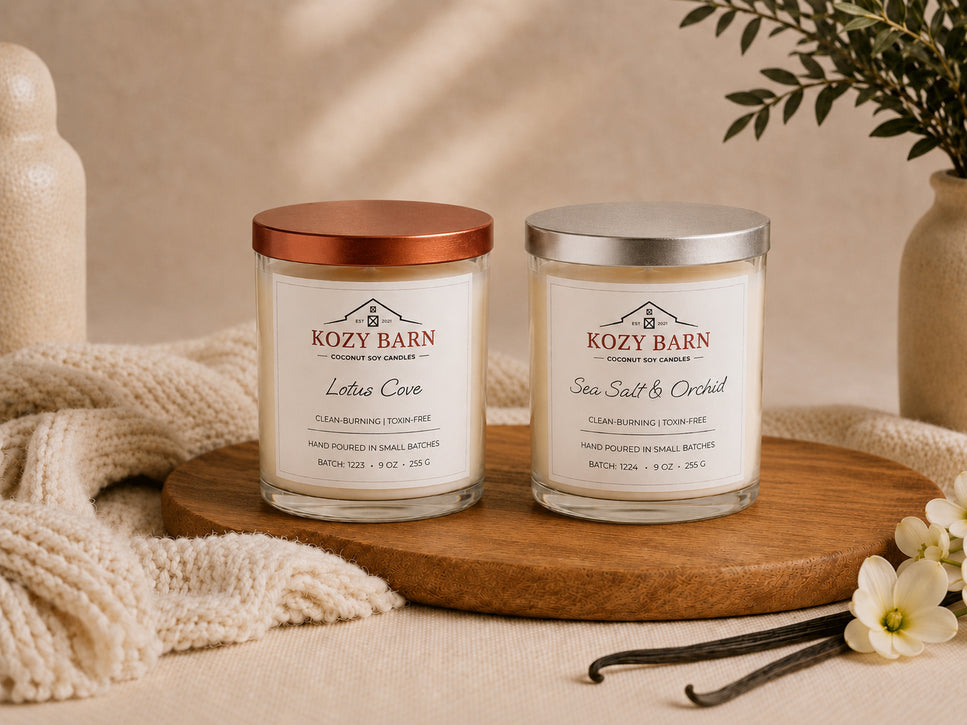 Kozy Barn candles one with copper or bronze lid labeled Lotus Cove and another with a silver lid labeled Sea Salt & Orchid. Both candles sitting on wooden display next to cozy knitted blanket and white blossom flowers. 