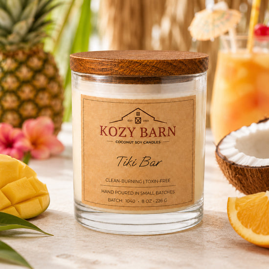 Kozy Barn candle with coconut soy wax and a wooden lid labeled Tiki Bar styled on a tabletop with tropical props including a pineapple, mango, orange slice, cocktail glass, and coconut half.
